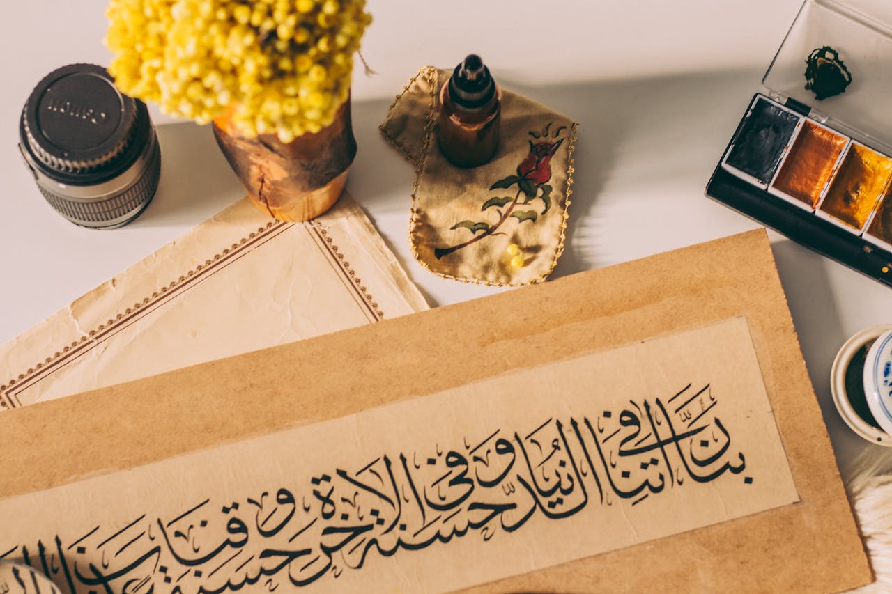 About Artistic setup featuring Arabic calligraphy, flowers, ink, and watercolor paints on a desk.